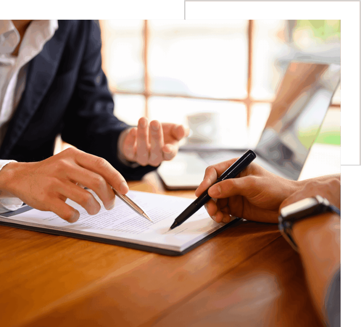 Two people in a business setting reviewing documents with pens on a wooden table near a laptop.