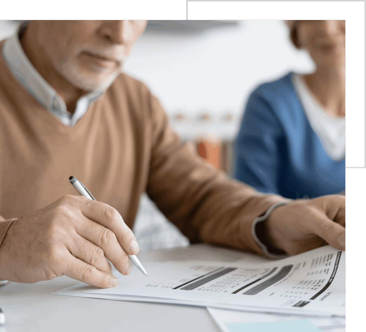 An elderly man reviewing documents and writing notes while a woman sits beside him in a home setting.