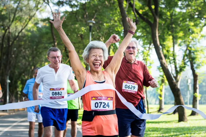 A joyful elderly woman crossing a finish line with her arms raised, accompanied by two elderly men running in a park.