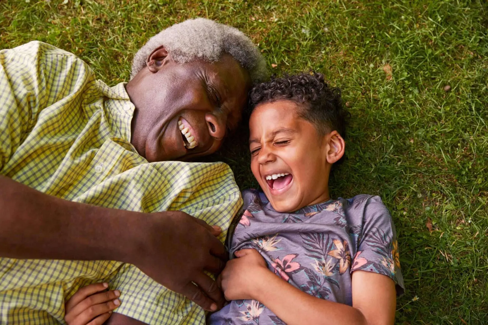An elderly man and a young boy lying on grass, sharing a joyful moment with big smiles and laughter.