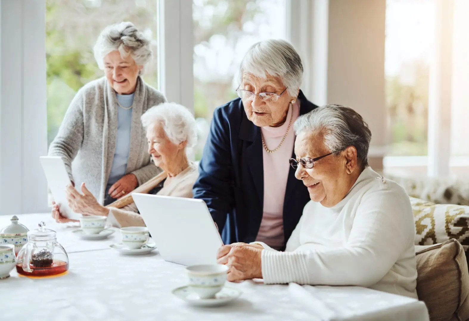 Four elderly women gathered around a laptop, smiling and engaging in a bright, cozy room.