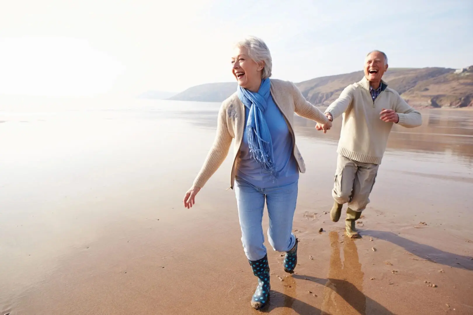An elderly couple joyfully running hand in hand along a sunny beach shore.