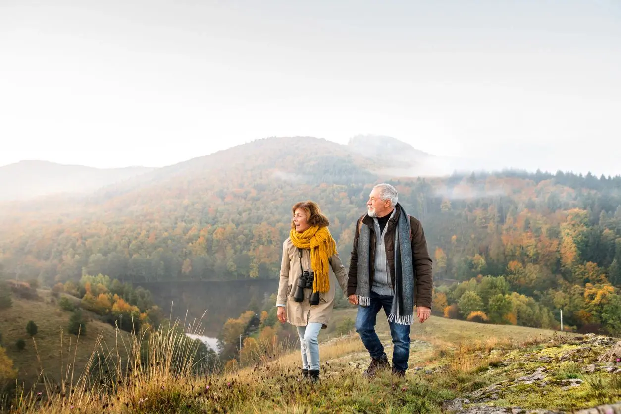 A couple dressed in warm clothes walking hand in hand on a grassy hill with foggy mountains in the background.