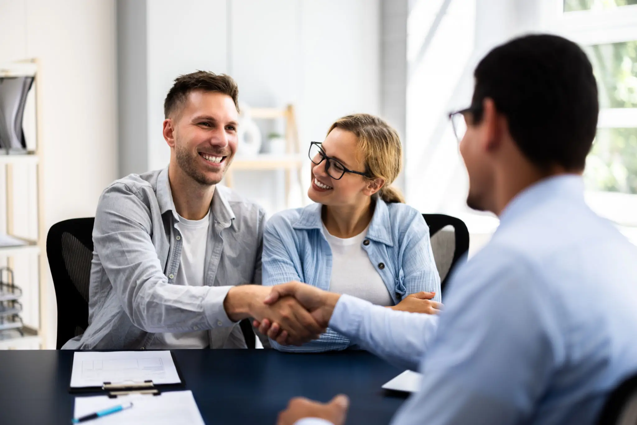 Two business professionals shaking hands while a colleague looks on during a meeting in a bright office.